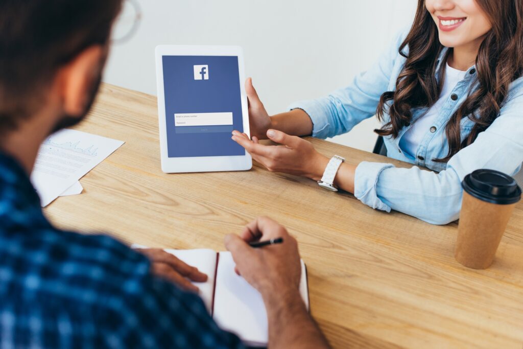 partial view of businesswoman with tablet with facebook logo and colleague with notebook taking part